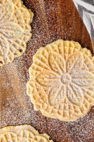 three pizzelli cookies with a dusting of powdered sugar and a white and black striped dishtowel