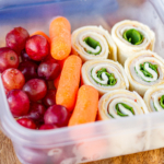 turkey hummus pinwheels in a plastic container with baby carrots and red grapes