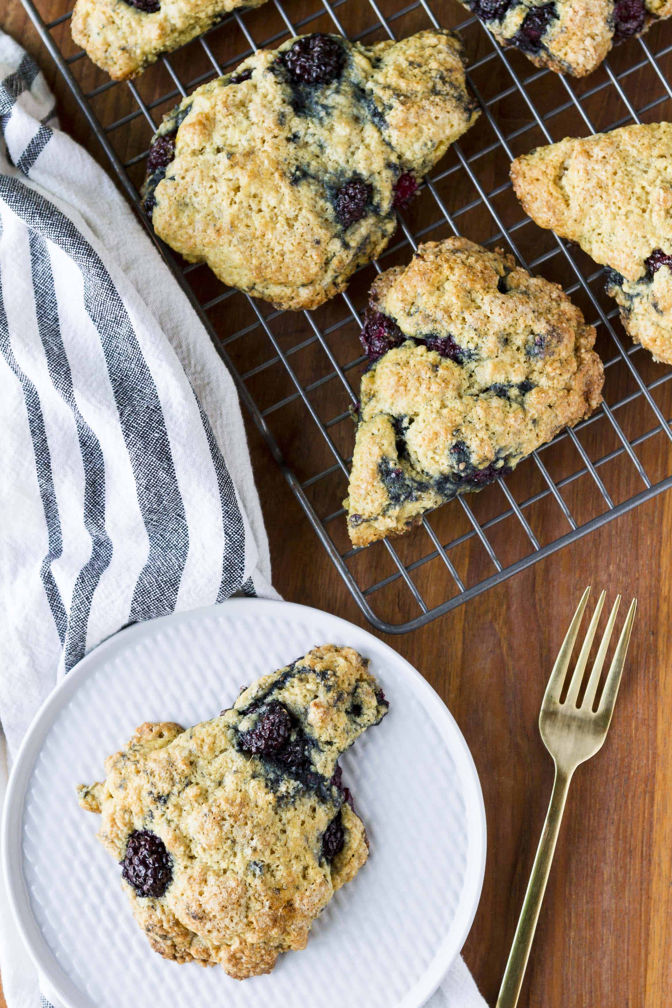 tender and buttery blackberry scones on a cooling rack with a single scone on a textured white plate with a gold fork next to it