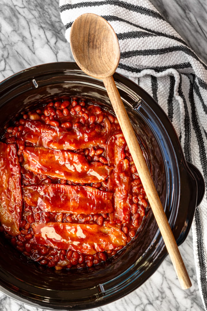 crockpot baked beans with bacon and brown sugar in a slow cooker insert with a wooden spoon