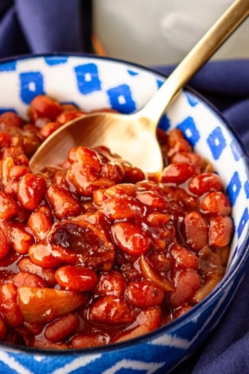 crock pot baked beans with bacon and brown sugar in a blue and white ceramic bowl with a gold spoon