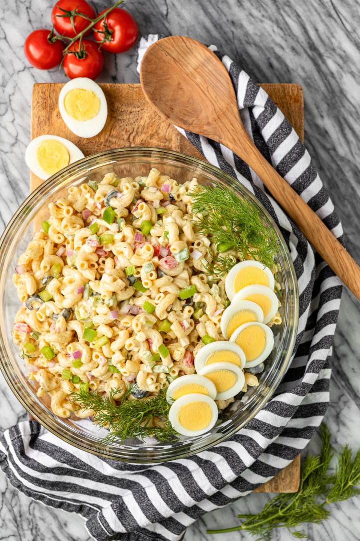 macaroni salad with egg in a glass bowl garnished with hard boiled eggs and fresh dill and a wooden spoon on the side
