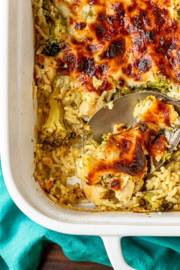 cheesy chicken and rice casserole recipe being scooped out of a white ceramic baking dish with a silver serving spoon