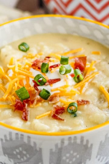 baked potato soup topped with sour cream, shredded cheddar cheese, crumbled bacon and sliced green onions in a ceramic bowl