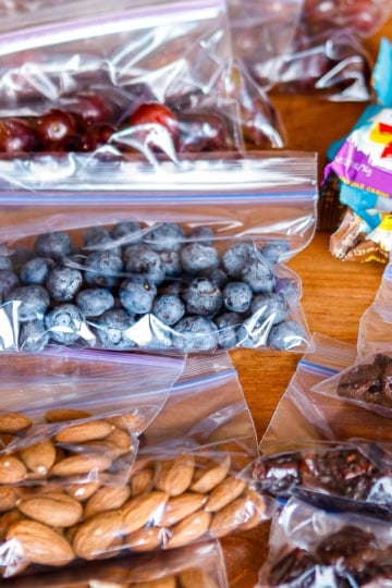 road trip snacks consisting of bagged blueberries, grapes, almonds, dried fruit, and granola bars on a wooden table