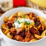 sausage chili recipe with beans in a white bowl topped with fritos corn chips, sour cream, shredded cheddar cheese, and chopped parsley