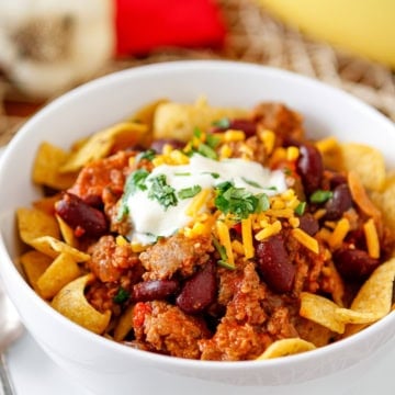 sausage chili recipe with beans in a white bowl topped with fritos corn chips, sour cream, shredded cheddar cheese, and chopped parsley
