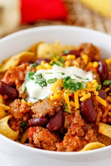 sausage chili recipe with beans in a white bowl topped with fritos corn chips, sour cream, shredded cheddar cheese, and chopped parsley